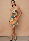 A woman modeling a sleeveless mini dress with a colorful floral print, standing against a textured beige wall.