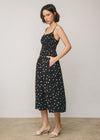 A woman modeling a black midi dress with white polka dots and thin spaghetti straps, standing against a neutral light gray studio background.
