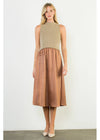 A woman modeling a sleeveless midi dress with a beige knit bodice and a brown faux-leather skirt, standing against a plain off-white wall.