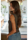 A woman with long brown hair is shown from the side/back wearing a sleeveless olive green top and light blue jeans in a bright, modern indoor setting.