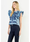 A woman modeling a light blue top with dark blue floral embroidery and ruffled sleeves, paired with dark jeans, against a plain light background.