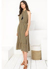 A woman modeling a sleeveless midi dress with a dense small-scale floral or paisley-like print in olive green, black, and beige, paired with brown strappy sandals, standing in a bright white studio setting.
