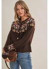 A woman wearing a dark brown knit cardigan with a colorful geometric Fair Isle pattern across the yoke and cuffs, paired with blue jeans and holding a brown leather crossbody bag against a solid light beige background.