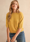 A woman wearing a mustard yellow knit sweater with elbow-length sleeves and blue jeans, standing against a textured beige wall.