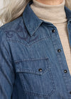 Blue denim jacket with embroidered design worn over a beige turtleneck sweater.