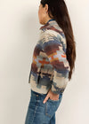 A woman shown from the side profile wearing a long-sleeve button-front jacket with a Southwestern geometric pattern in beige, blue, brown, and orange, paired with blue jeans, against a plain off-white background.