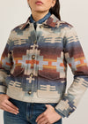 A woman wearing a cropped, long-sleeve button-front jacket with a Southwestern geometric pattern in beige, blue, brown, and orange, paired with a blue denim shirt collar and blue jeans.