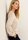 A woman with blonde hair leans against a white wall, wearing a light beige knit sweater with red trim and dark navy pants.