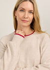 A woman wearing a light beige knit sweater with red ribbed trim around the crew neck and cuffs, photographed against a plain light gray background.