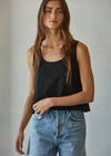 A woman wearing a black sleeveless tank top and light blue denim jeans, standing against a plain light gray wall.