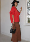 Woman in red sweater and brown skirt holding a black handbag indoors.