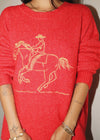 Red sweater with a white graphic of a person riding a horse worn by a person.