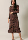 A woman modeling a long-sleeve midi dress with a sheer brown mesh fabric and a black floral lace overlay, set against a plain light beige studio background.