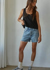 A woman modeling a black sleeveless tank top and light-wash denim shorts with frayed hems, paired with white socks and gray sneakers, standing in a minimalist room with a white curtain on the left.