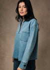 A woman wearing a light blue denim shirt jacket with a half-button placket and chest pocket, paired with dark jeans, posed against a textured brown studio backdrop.
