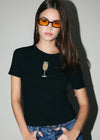 Person wearing a black t-shirt with a wine glass design and orange sunglasses against a plain background