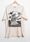 Beige t-shirt with a graphic of a woman in a dress and stars on a hanger against a white brick wall.