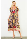 A woman modeling a short-sleeve midi dress with a dark brown base and a colorful floral and scroll pattern in pink, red, yellow, and beige, standing against a plain white wall.