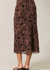 A model's lower body wearing a brown midi skirt with a black floral vine pattern, paired with black slide sandals, against a plain light beige background.