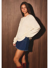 A woman posing in a cream-colored knit crewneck sweater, a blue denim mini skirt, and brown boots against a dark wood-paneled wall.