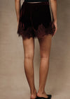 A person shown from the back wearing dark burgundy velvet shorts with a lace hem, paired with black mule heels, against a plain beige studio backdrop.