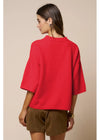 A woman shown from the back wearing a bright red knit sweater with wide three-quarter sleeves and brown shorts, set against a plain beige studio background.