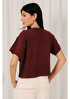 A woman shown from the back wearing a short-sleeve textured knit sweater in deep burgundy, paired with off-white pants, set against a soft white curtain background.
