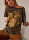 A woman wearing a dark olive green off-the-shoulder t-shirt with a large yellow rose graphic on the front, paired with brown pants, leaning against a neutral wall.