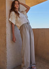 Woman in a white knit top and beige pants leaning against a textured wall with a clear sky.