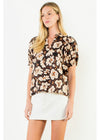 A blonde model wearing a short-sleeve button-up blouse with a brown, beige, and white floral pattern, paired with a white mini skirt, against a plain white studio background.