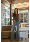 A woman modeling a sleeveless olive-brown top and light-wash blue flare jeans in a modern indoor setting with large windows, wooden ceiling panels, and a tan armchair.