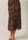 A brown midi skirt with a black floral pattern, shown on a model from the waist down wearing black strappy heels against a light beige studio background.