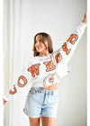 Woman wearing a white long-sleeve shirt with 'Cowboys' printed in orange, standing against a white curtain.