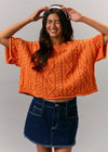 Woman wearing an orange knitted sweater and denim skirt against a plain background