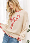 Woman wearing a cream sweater with a red lobster design, standing indoors.