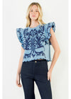 A woman modeling a light blue top with a dark blue floral damask pattern and ruffled sleeves, paired with dark jeans, against a plain light gray background.