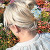 Person with blonde hair tied back with a floral hair accessory, standing in front of a colorful flower background.
