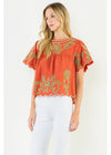A woman modeling a short-sleeve orange blouse with green and white embroidered patterns and a scalloped hem, paired with white pants, against a plain white background.