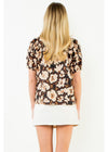 A woman viewed from behind wearing a short-sleeve floral blouse with a dark brown base and beige, cream, and orange flowers, paired with a white skirt, against a plain white background.