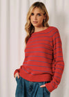 A woman wearing a red knit sweater with thin blue horizontal stripes, paired with blue jeans, standing against a white curtain background.