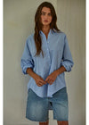 A woman wearing a light blue and white vertically striped button-down shirt with rolled-up sleeves and light-wash denim shorts, standing against a textured off-white wall.