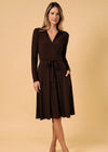 A woman modeling a dark brown long-sleeve wrap-style dress with a tie waist and side pockets, standing against a solid beige studio background.