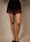 Black velvet shorts with a burgundy lace hem, modeled with black pointed-toe heels against a neutral beige studio backdrop.