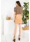 A woman is shown from behind wearing a brown and beige floral patterned puff-sleeve top and a tan faux-leather mini skirt, paired with tan ankle boots, in a bright white room with a potted plant.