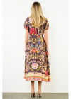 A woman viewed from behind wearing a short-sleeve midi dress with a brown base and a colorful floral and scroll pattern, paired with beige high-heeled sandals against a plain white studio background.