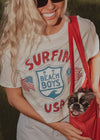 Person wearing a 'Surfin' Beach Boys USA' t-shirt holding a small dog in a red bag.