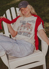 Person sitting on a white chair wearing a graphic t-shirt and red scarf, with a casual outdoor setting.