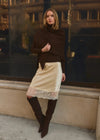 A woman stands outdoors in front of a window wearing a dark brown wrap, a cream satin pencil skirt with a lace hem, and dark brown over-the-knee boots.