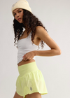 A woman modeling a white ribbed tank top and a light yellow-green athletic skirt with a small embroidered logo, paired with a cream beanie, against a plain light gray studio background.