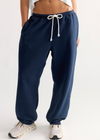 A person wearing navy blue jogger sweatpants with a white drawstring, paired with a white cropped top and light-colored sneakers, shown from the waist down against a plain light background.
